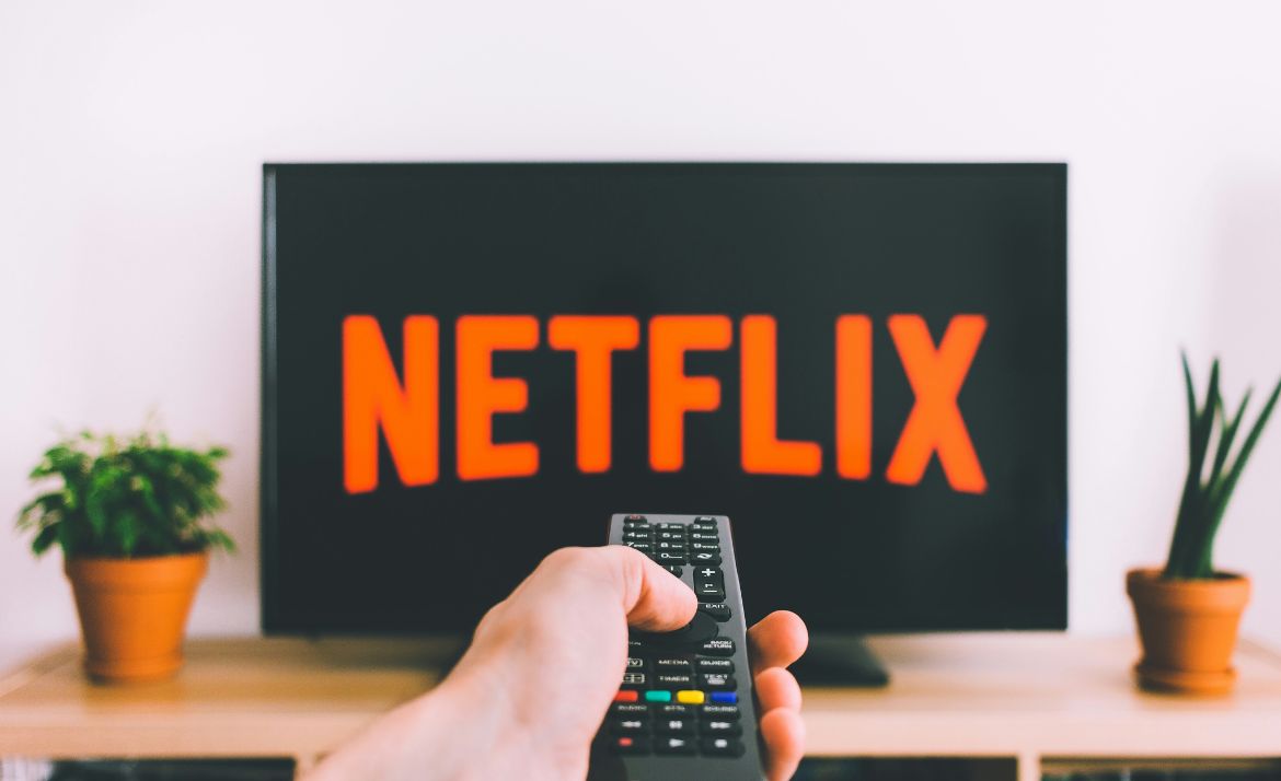 Hand hält Fernbedienung und richtet sie auf Fernseher mit dem Schriftzug 'NETFLIX'