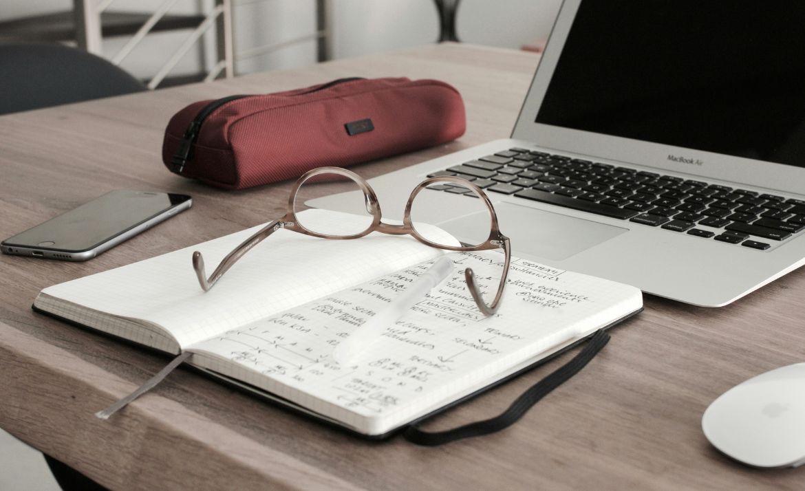 Arbeitsplatz mit offenem Notizbuch, Brille, Laptop, Smartphone und rotem Stifteetui auf Holztisch