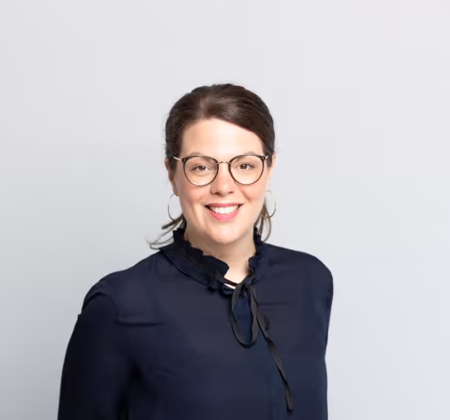 Porträt einer lächelnden Frau mit dunklen Haaren, die eine Brille, goldene Creolen und eine dunkelblaue Bluse trägt.
