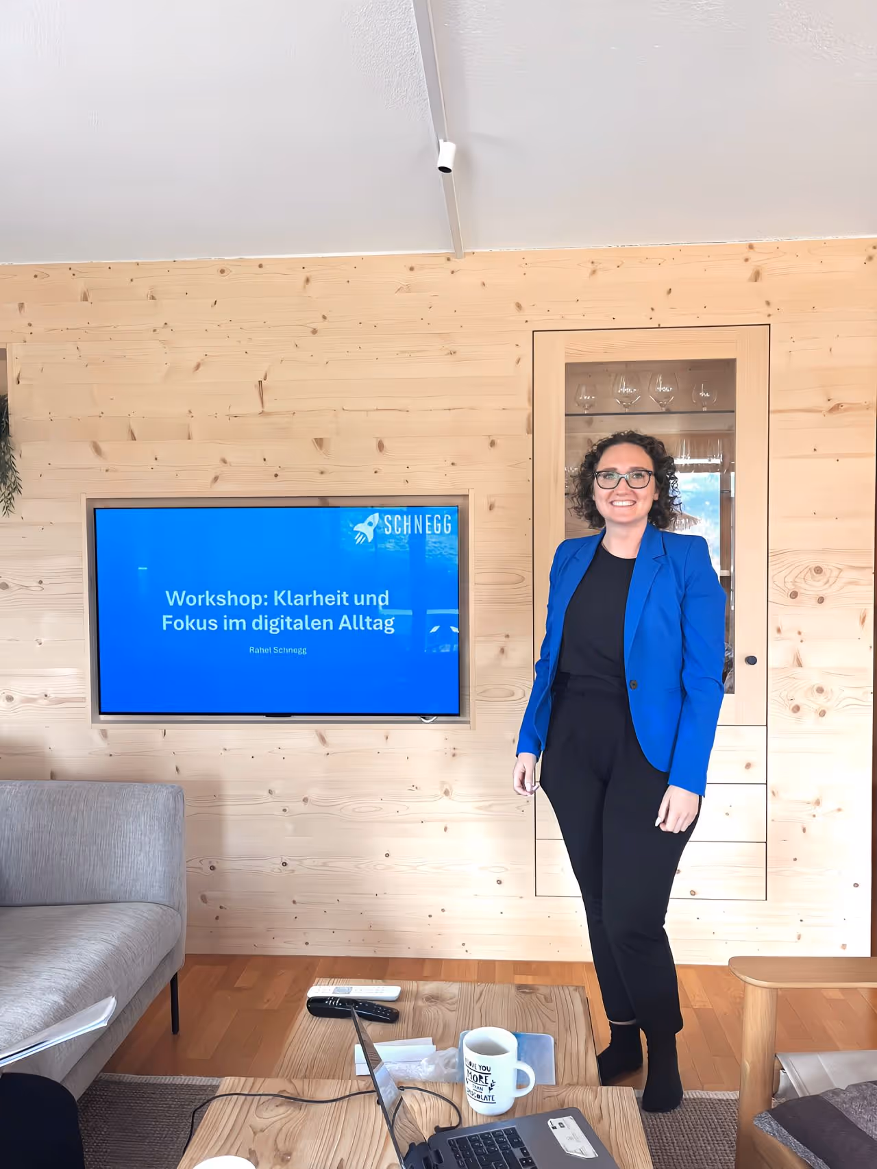Frau mit blauer Jacke und schwarzer Kleidung steht im modernen Raum mit Holzpaneelen, im Hintergrund Bildschirm mit Titel eines Workshops über Klarheit und Fokus im digitalen Alltag.