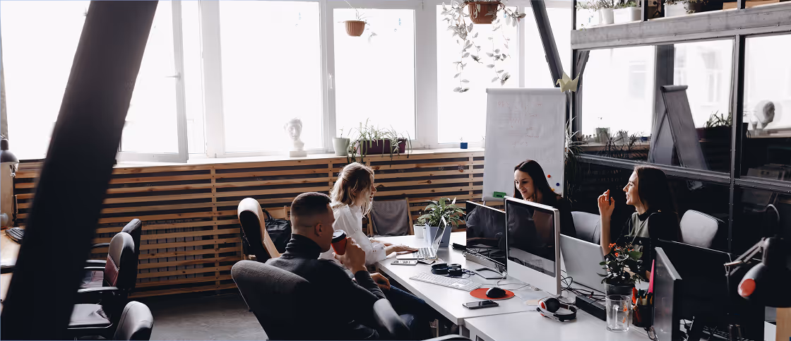 Quatre personnes travaillant sur des ordinateurs portables et écrans dans un bureau moderne avec lumière naturelle et plantes.