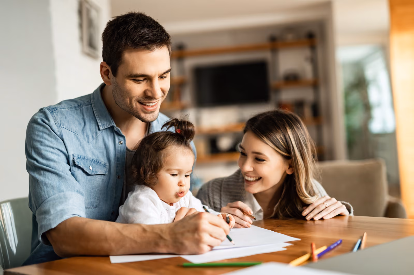 A mother, father and infant are happily spending time together drawing on a piece of paper at a wooden table inside their home.