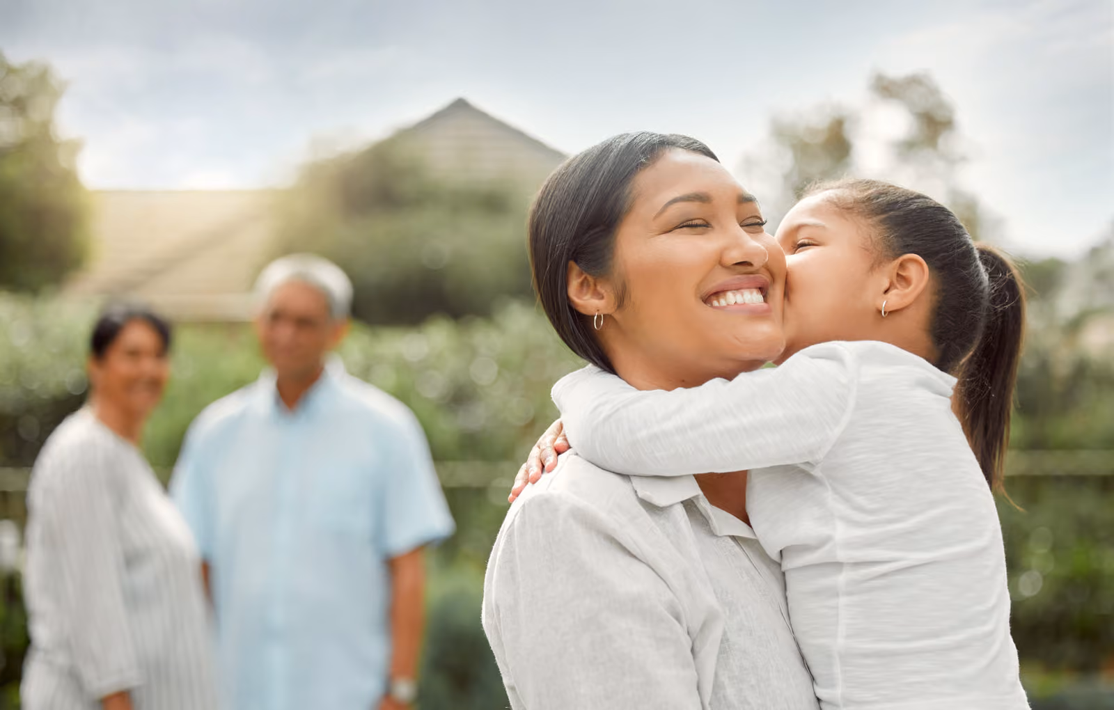 A woman with short dark hair hugs a child, with a home and greenery in the background.