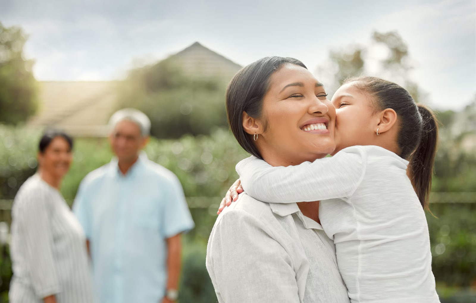 A woman with short dark hair hugs a child, with a home and greenery in the background.