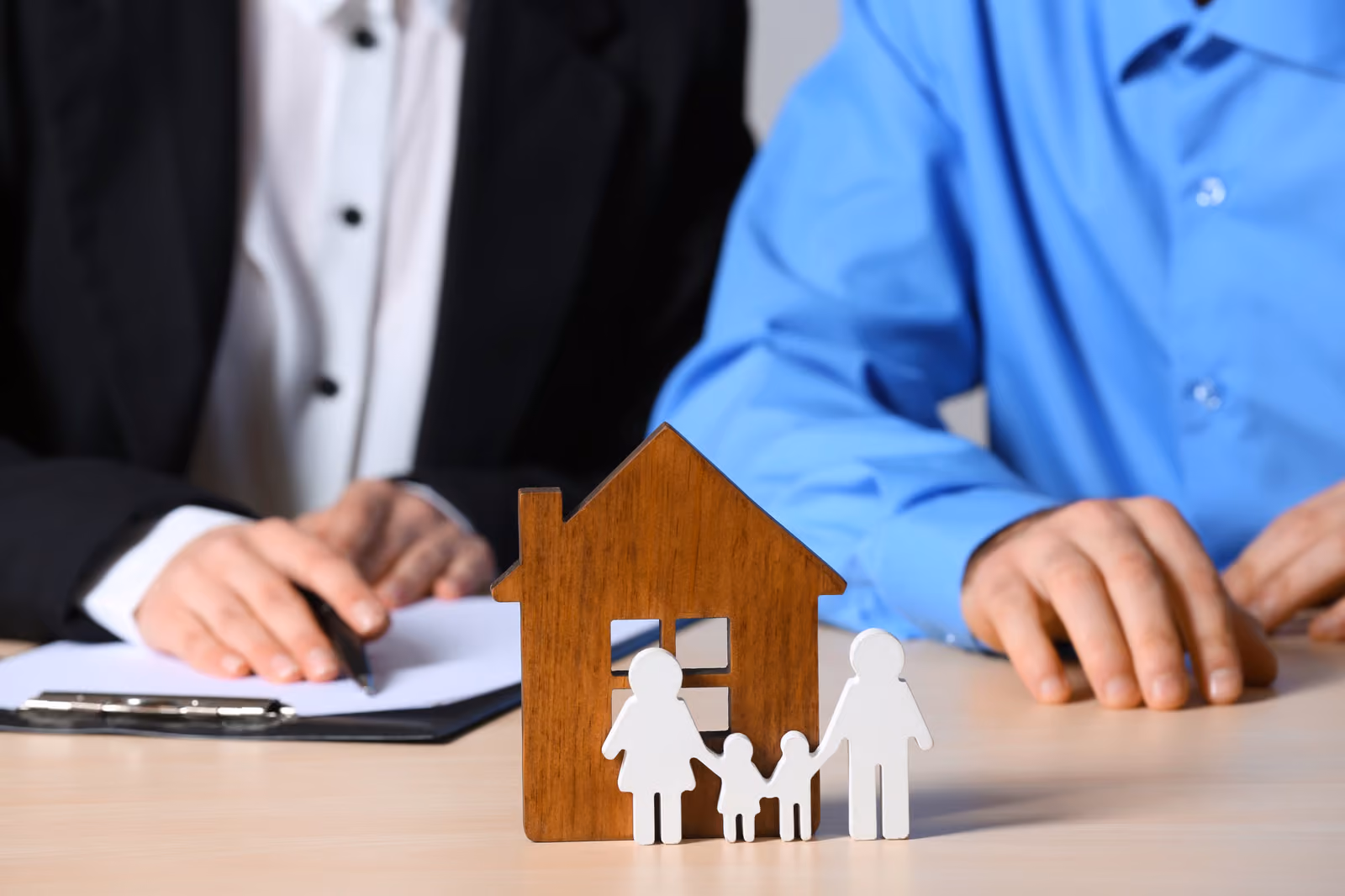 Two adults are partially visible sitting at a table with a miniature wooden house and a family of figurines.