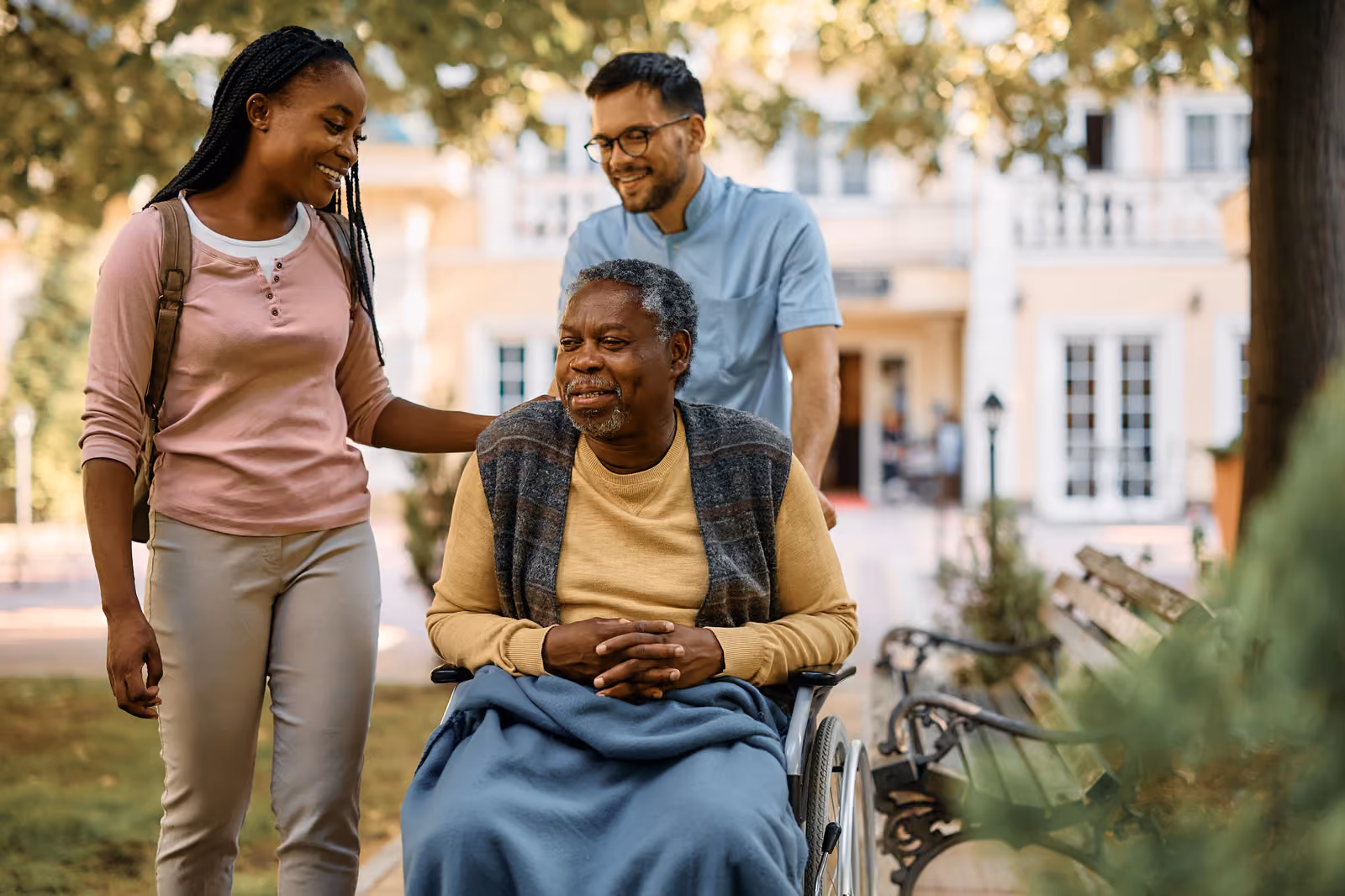 A young woman and a man are walking with a senior man in a wheelchair through a leafy park.