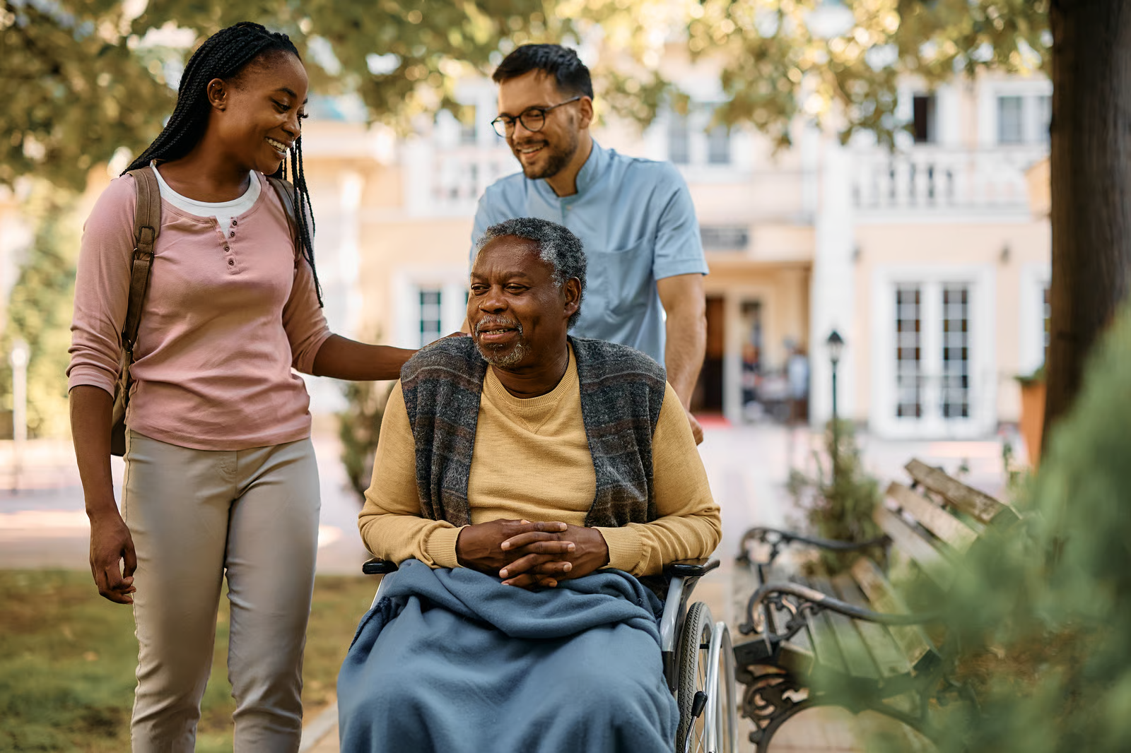 A young woman and a man are walking with a senior man in a wheelchair through a leafy park.