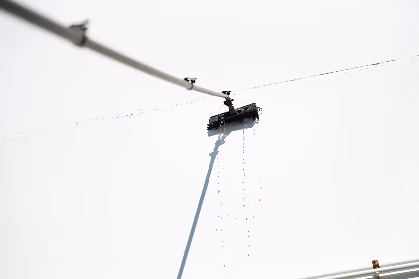 Automated window cleaning robot suspended on cables, spraying water while cleaning a glass surface.