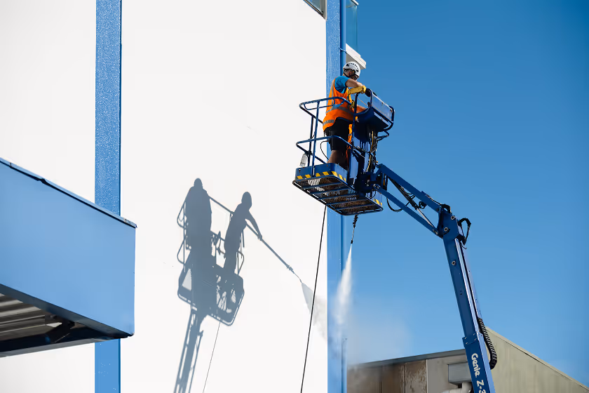 Worker in safety gear pressure washing a white building facade from an elevated hydraulic lift under clear blue sky.