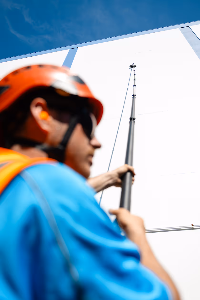 Worker in an orange helmet and blue shirt holding a tall pole against a white wall under a blue sky.