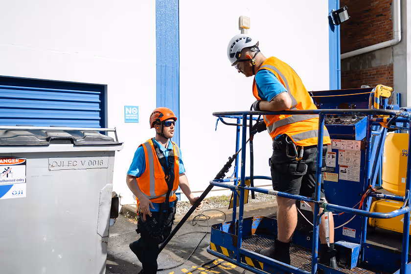 Two construction workers wearing helmets and orange safety vests operating a blue scissor lift near a building.