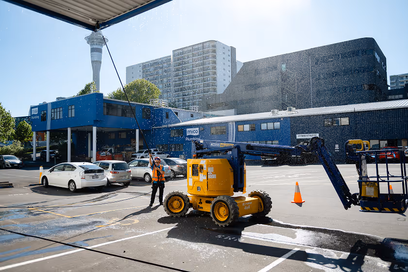 Person in safety vest using a hose to spray water on a large yellow lift machine in a parking lot under bright daylight.