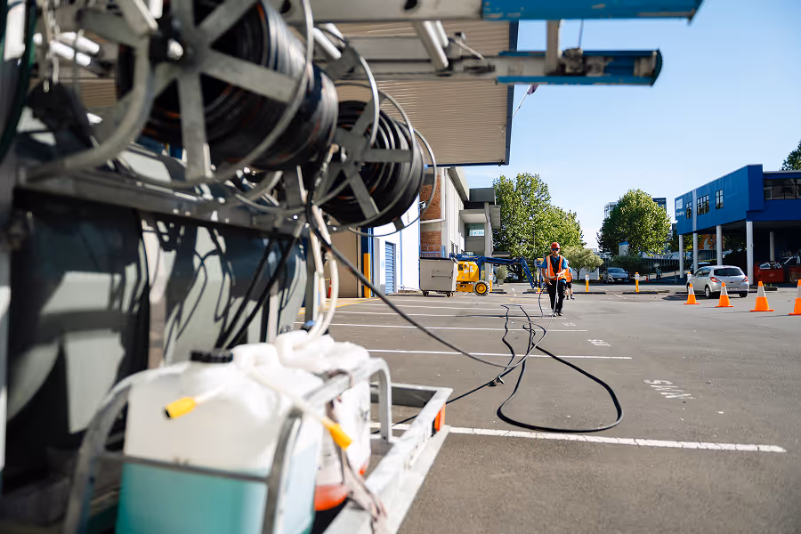Worker in safety vest and helmet using hose reels for power washing in an outdoor parking lot with buildings and traffic cones.