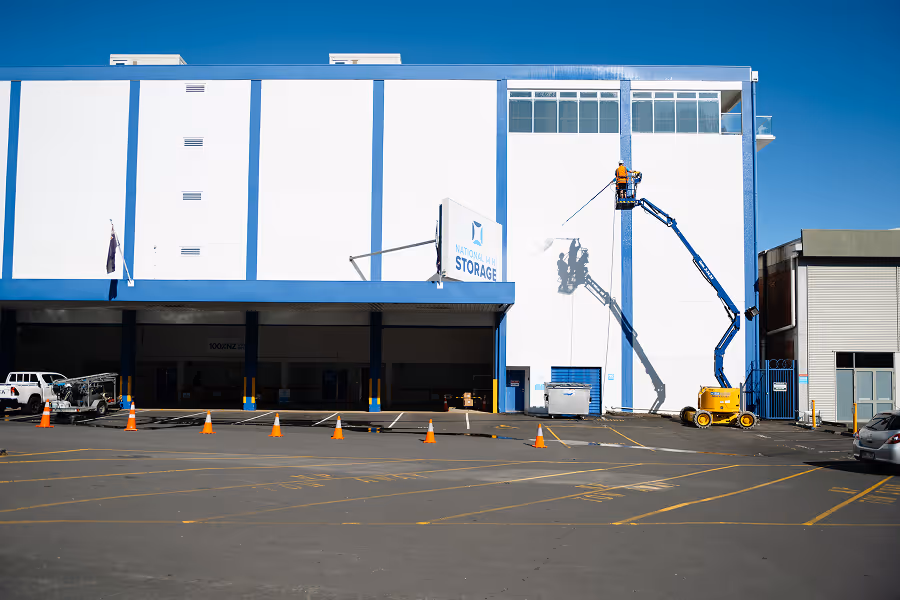 A worker in an orange jacket on a yellow lift painting the upper side of a white and blue storage building with a clear blue sky background.
