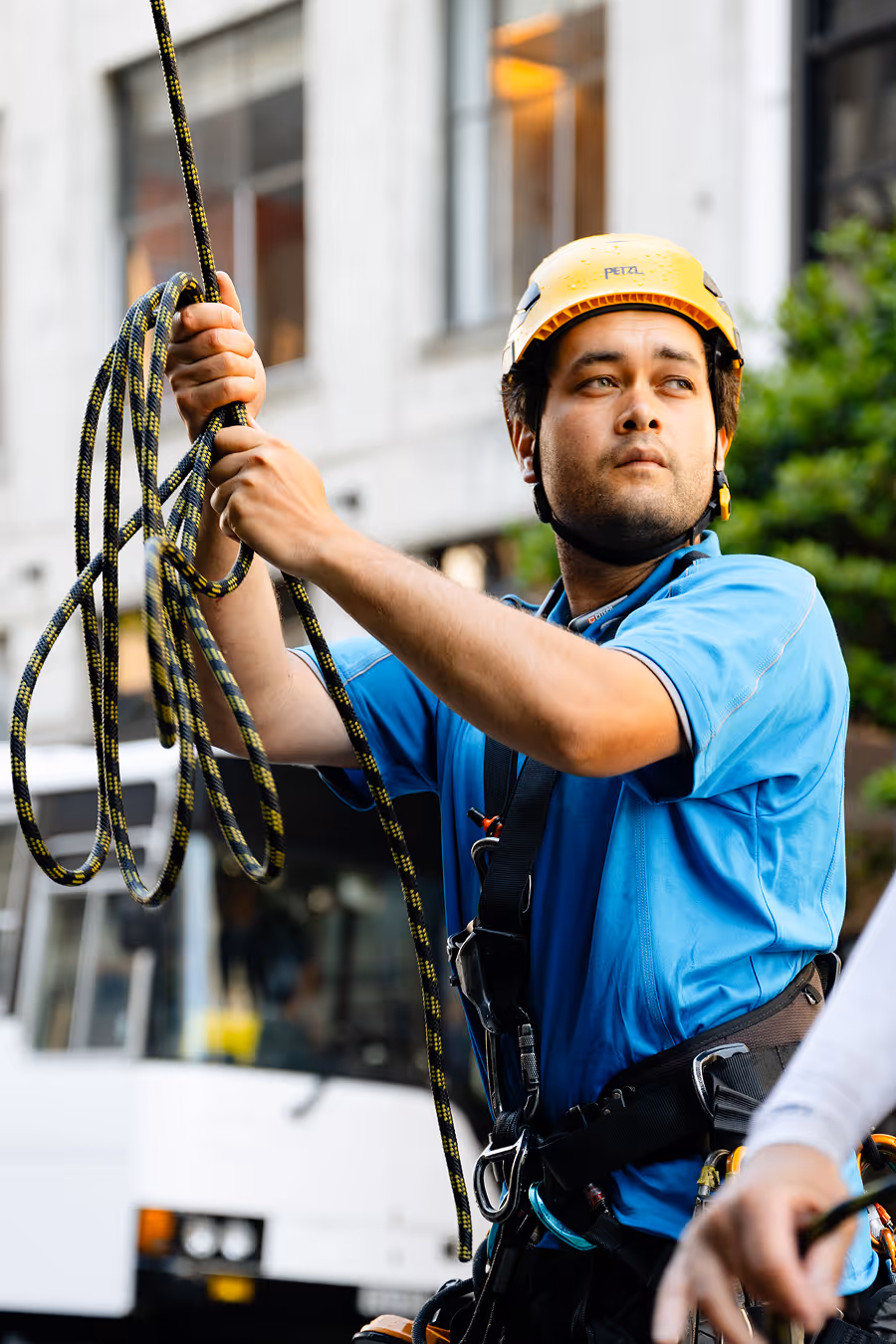 Worker in a yellow safety helmet and blue shirt holding climbing rope, preparing for work outdoors.
