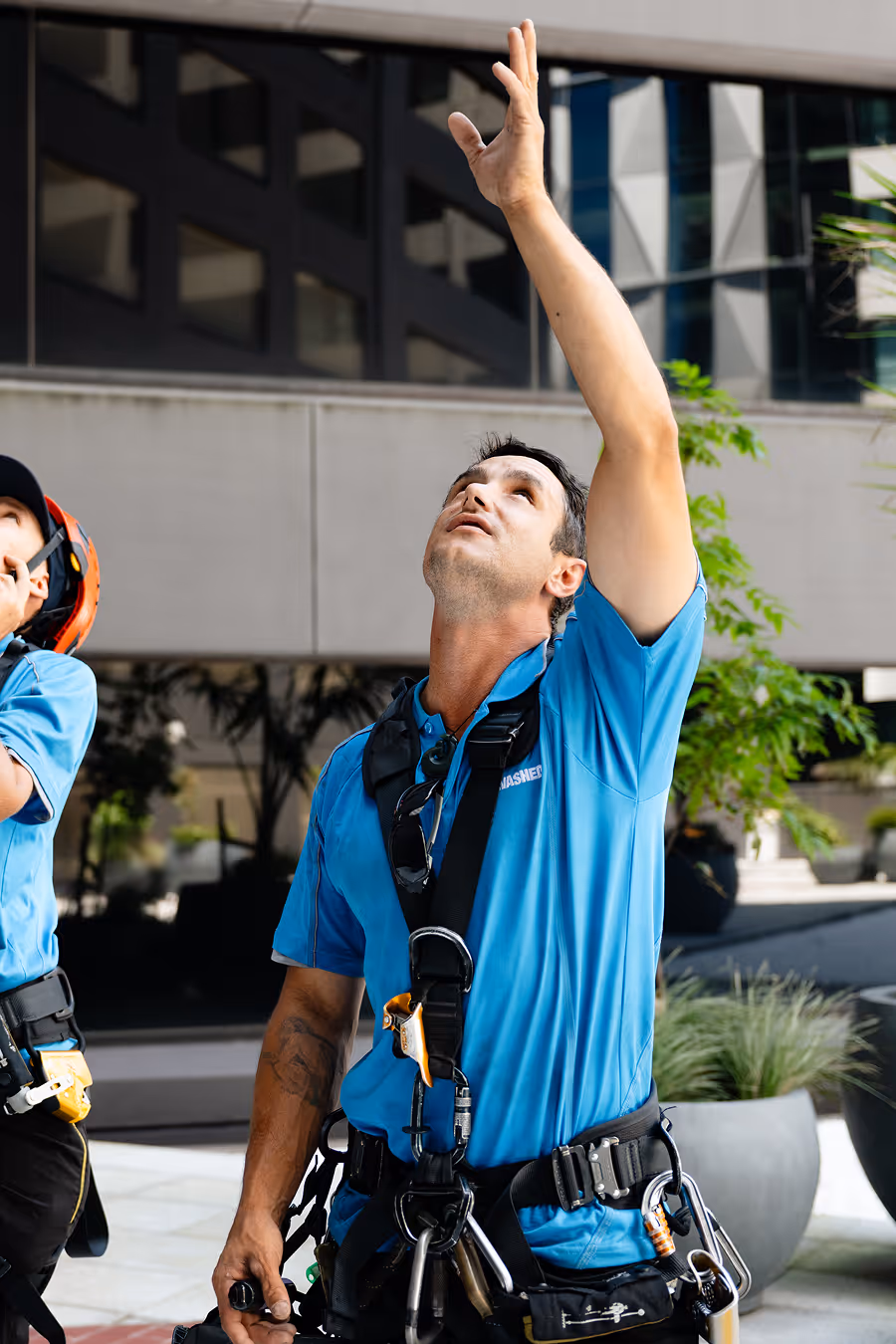 A worker in a blue shirt wearing a safety harness is looking up and reaching his hand upwards near a building.