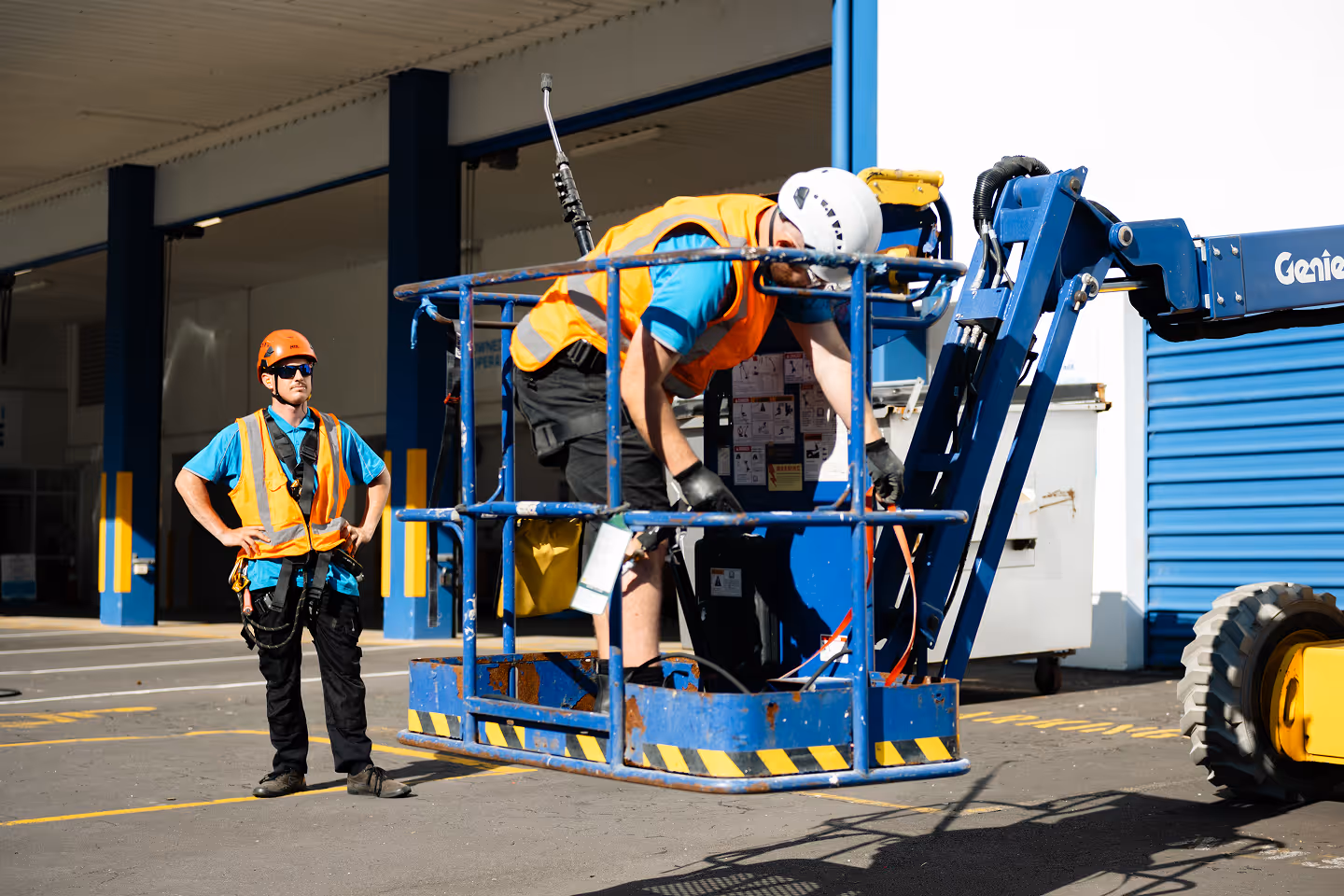 Two construction workers in safety gear operating and monitoring a blue Genie aerial lift outside a building.