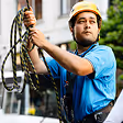 Construction worker wearing a yellow helmet and blue shirt holding a rope at a worksite.