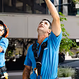 Two men in blue uniforms working outdoors, one looking up while reaching with his right arm.
