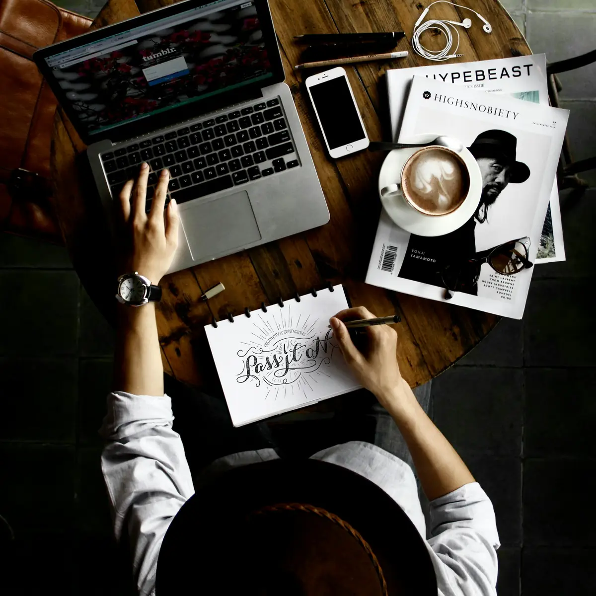 Designer sketching ideas in a notebook next to a laptop, phone and coffee while developing a new service