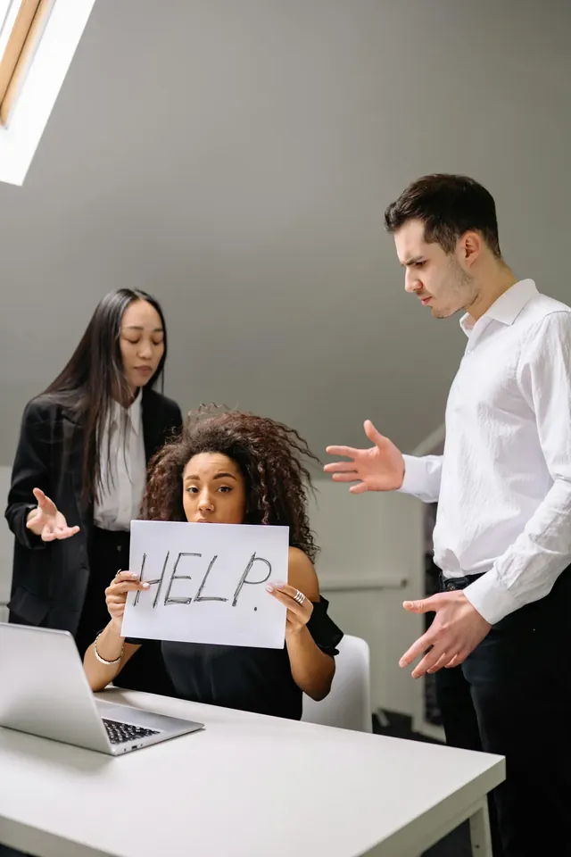 Konflikt im Büro: Zwei Kollegen diskutieren gestikulierend über eine sitzende Kollegin hinweg, die ein Schild mit der Aufschrift Help hochhält.