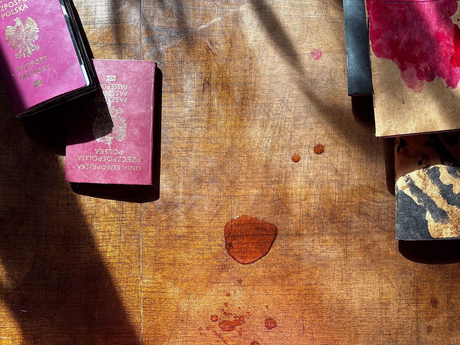 Wooden table in sunlight with two Polish passports, books and small liquid stains.