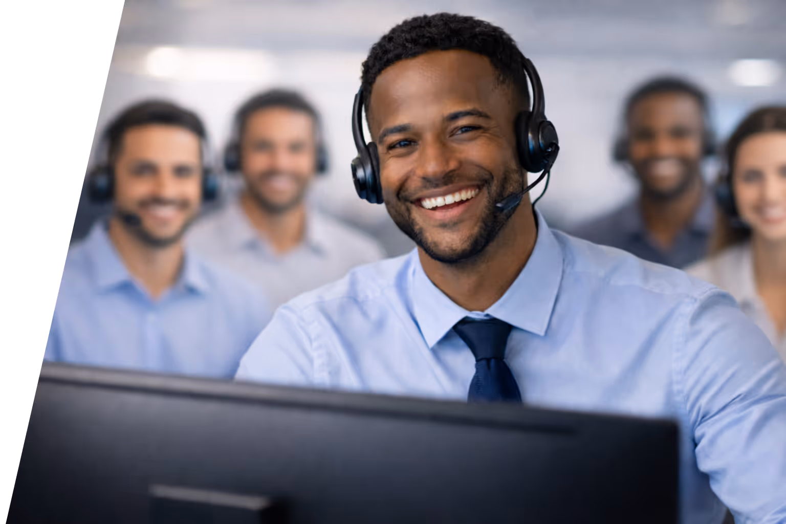 Smiling customer service representative wearing headset in front of a computer with colleagues in the background.