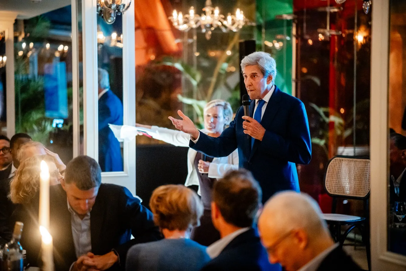 Man in a blue suit holding a microphone and speaking to an audience in a warmly lit indoor setting.
