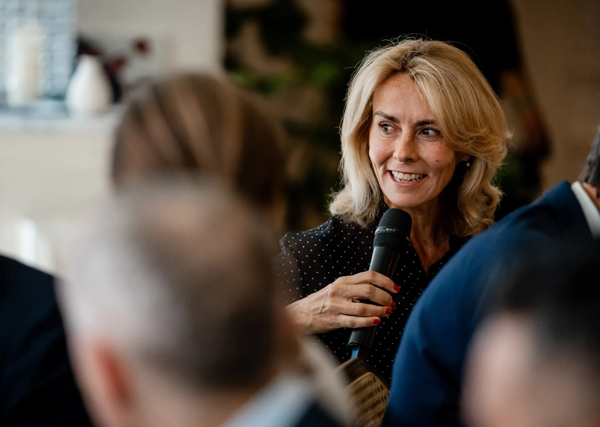 Smiling blonde woman speaking into a microphone at an indoor event.