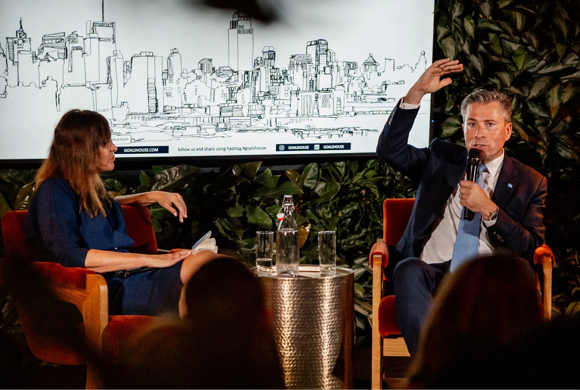 Man in a suit speaking into a microphone with his hand raised, seated next to a woman listening attentively, both engaged in a discussion on stage.
