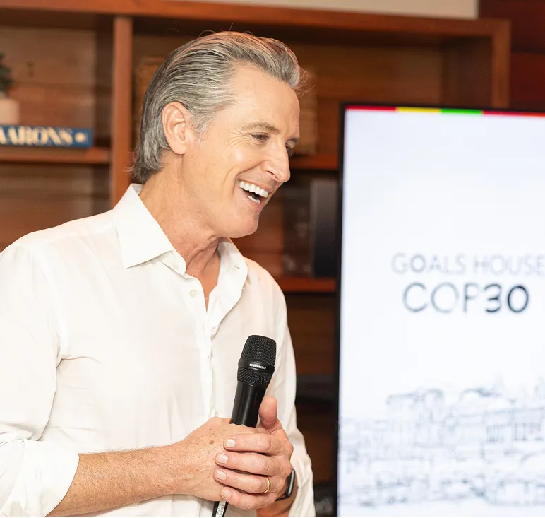 Smiling man in a white shirt holding a microphone while standing in front of a screen displaying 'GOALS HOUSE COP30'.