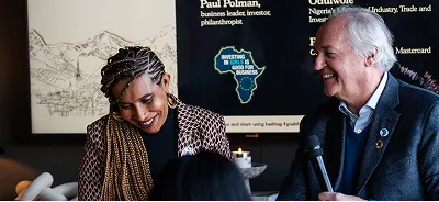 Two people smiling and engaged in conversation, one holding a microphone, with a business event backdrop behind them.