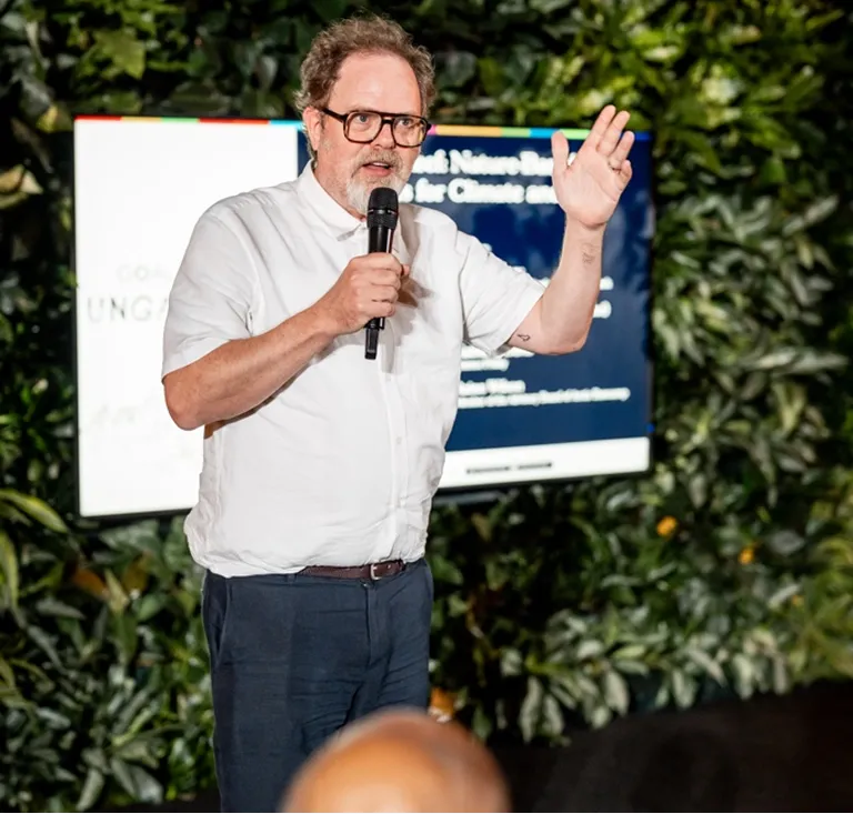 Man with glasses speaking into a microphone and gesturing with his hand during an outdoor presentation.