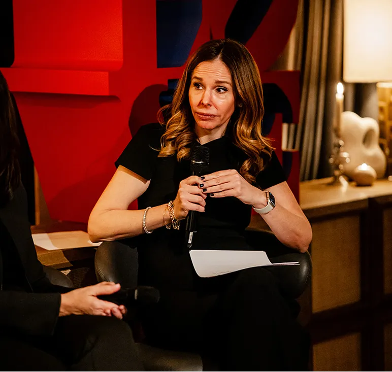 Woman with wavy hair holding a microphone and a paper, sitting in a chair during a discussion.