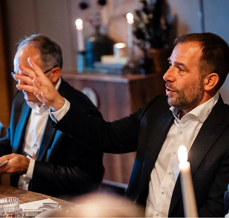 A man in a dark suit and white shirt gestures with his hand while speaking at a dinner table with lit candles.