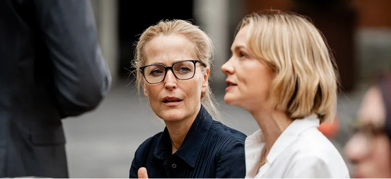 Two women engaged in conversation, one wearing glasses and a dark shirt, the other with short blonde hair in a white blouse.