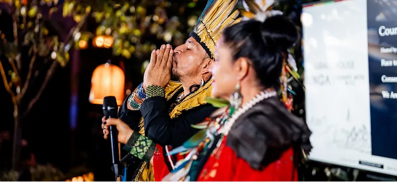 Two people in colorful traditional clothing, one speaking into a microphone and the other with hands clasped near their face.