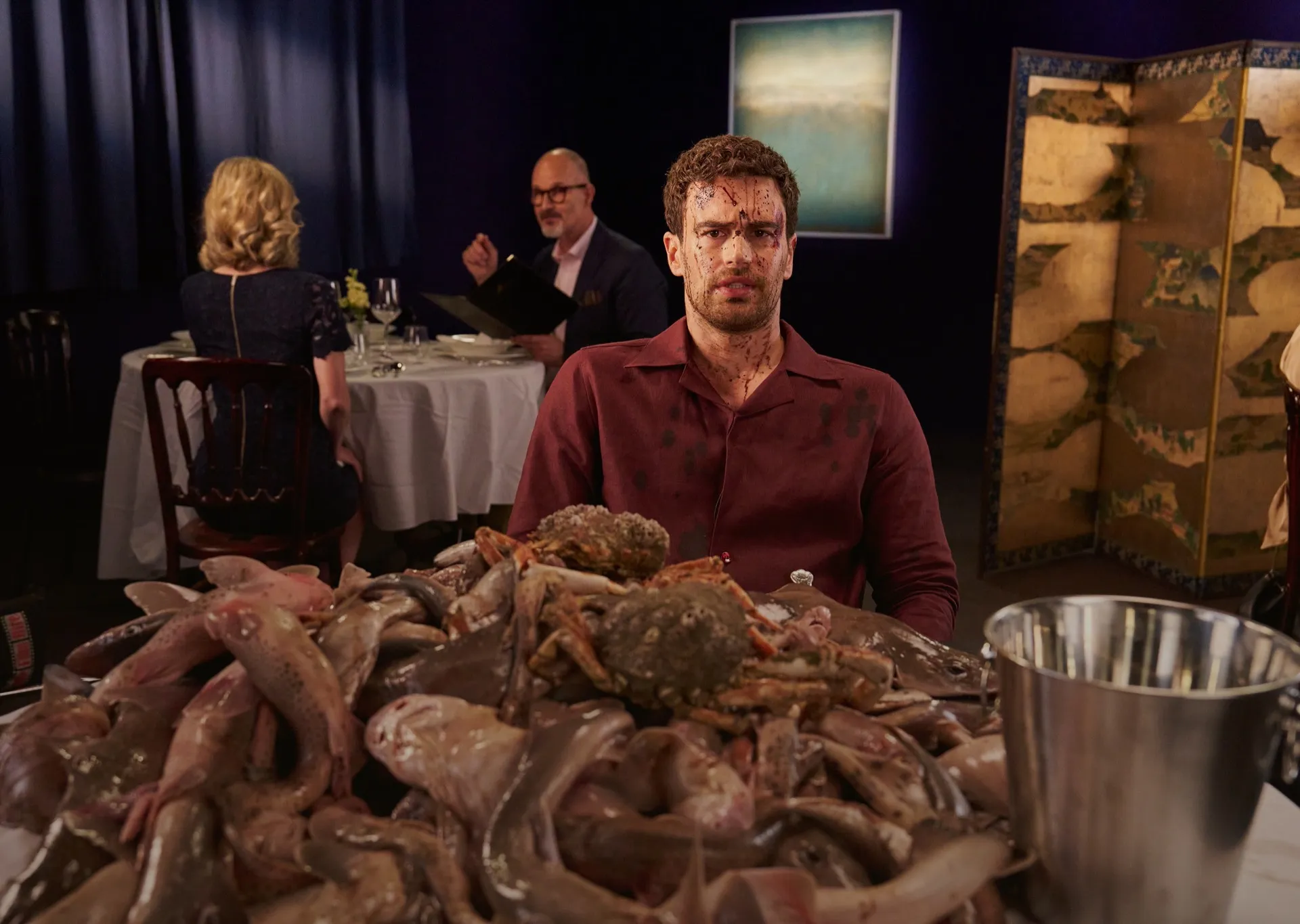 Man with blood on his face and shirt sits at a table piled with raw fish and crabs in a dimly lit restaurant setting.