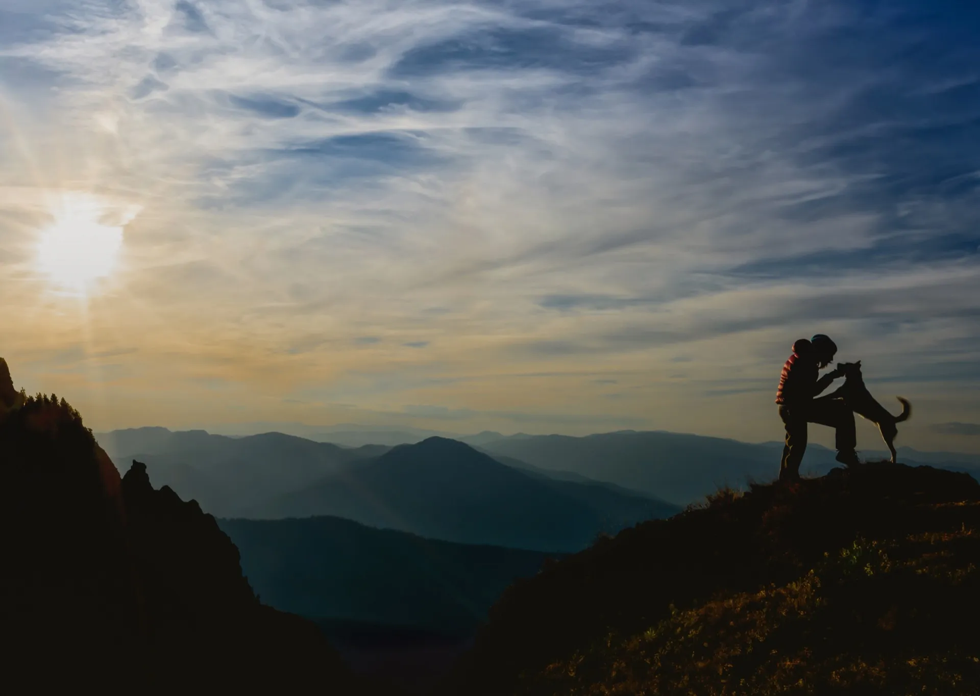 Silhouette of a person playing with a dog on a hilltop at sunset with mountains and a partly cloudy sky in the background.