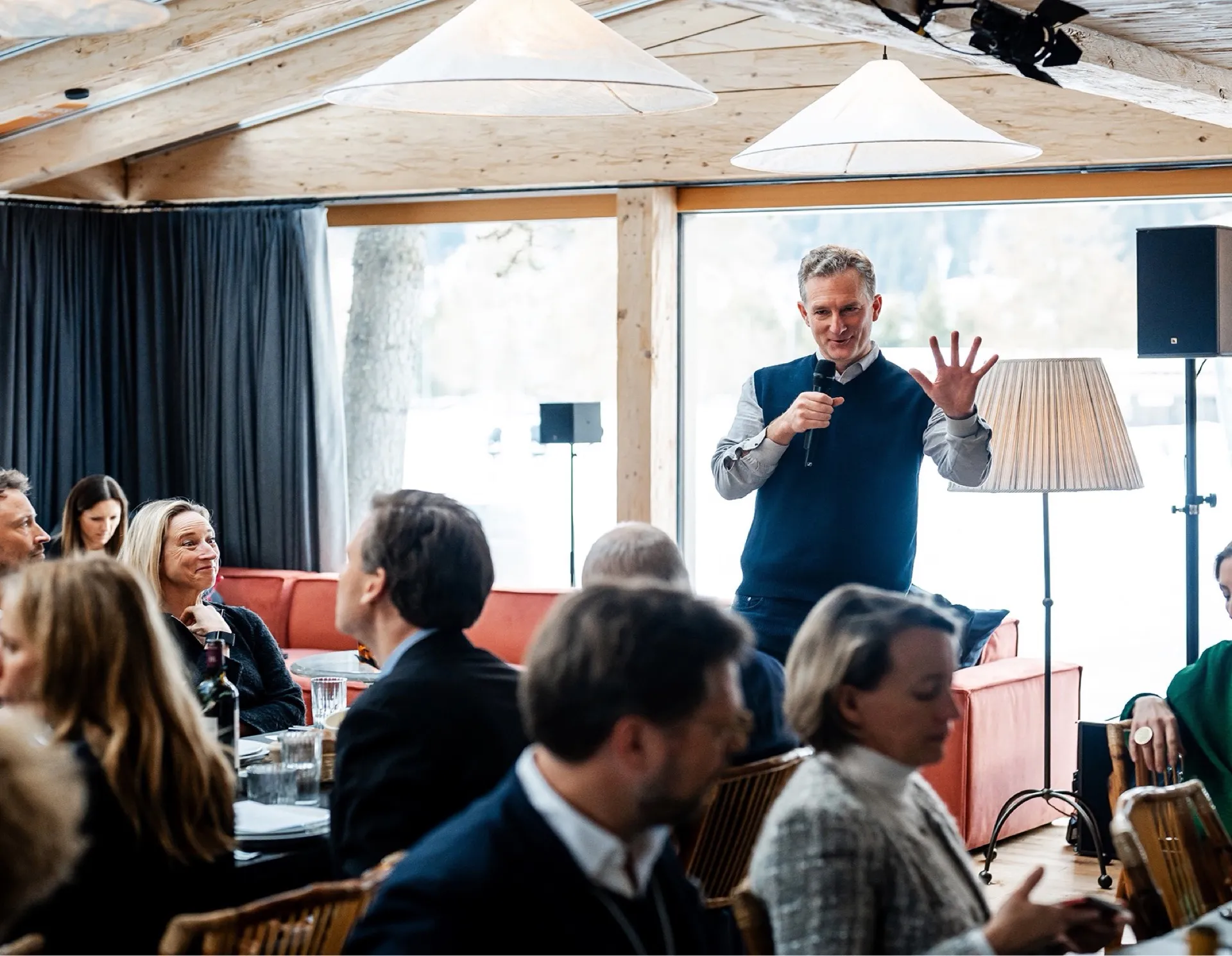 Man in a blue vest speaking into a microphone, gesturing with his hand, in a room with seated audience.