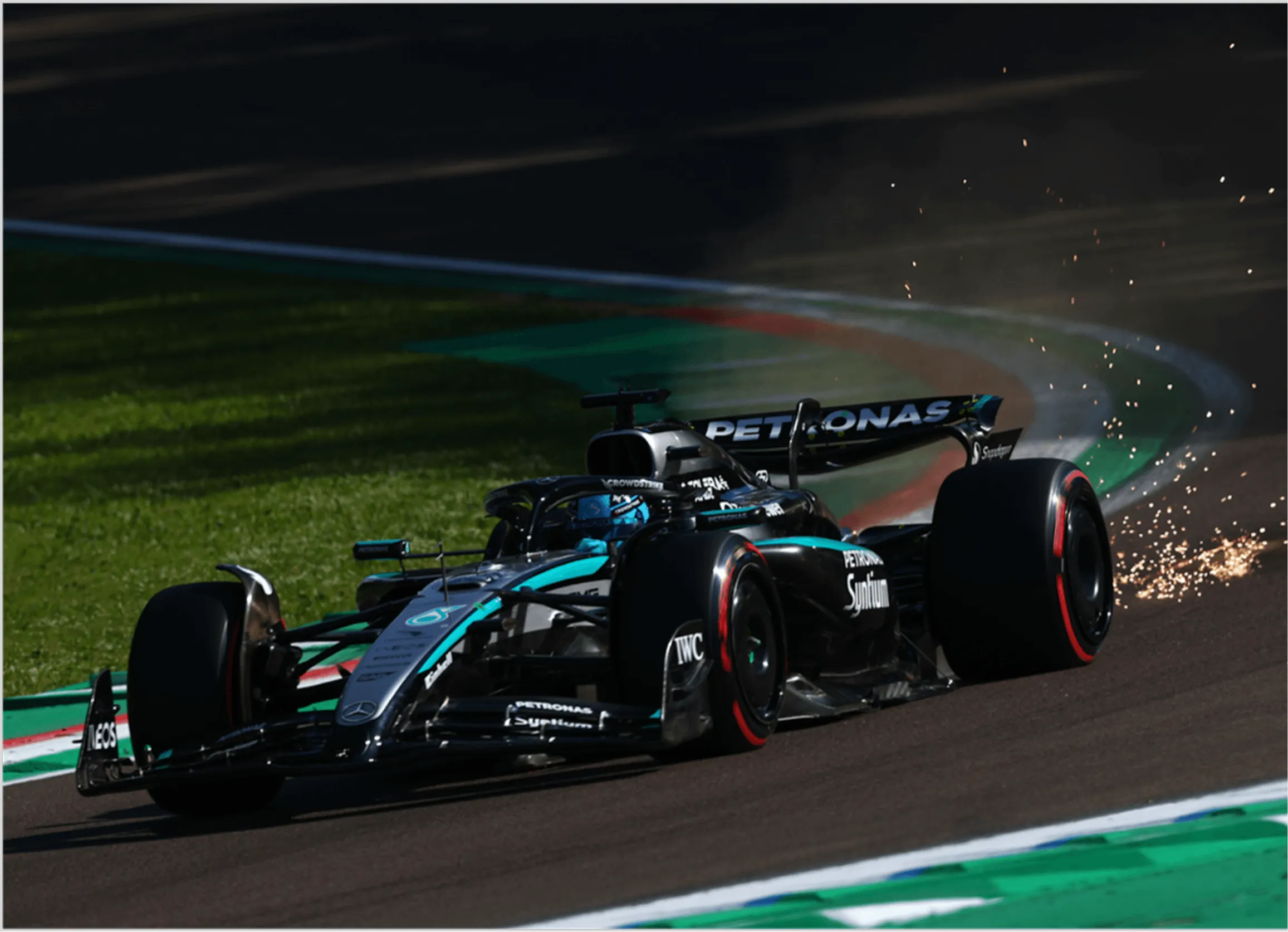 Mercedes Formula 1 car racing on track with sparks flying from rear right tire near a curve.