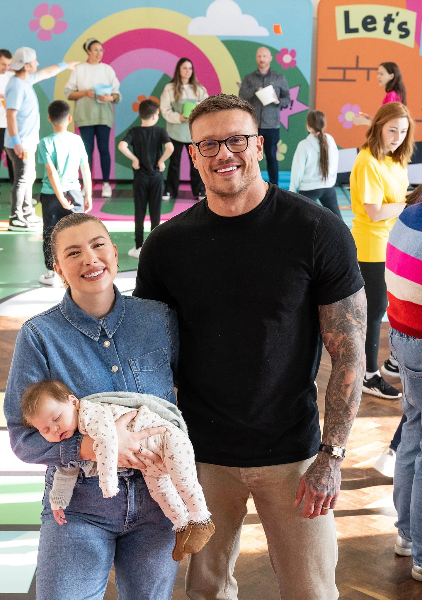 Smiling young couple with the woman holding a sleeping baby, standing in a colorful indoor play area with other children and adults.