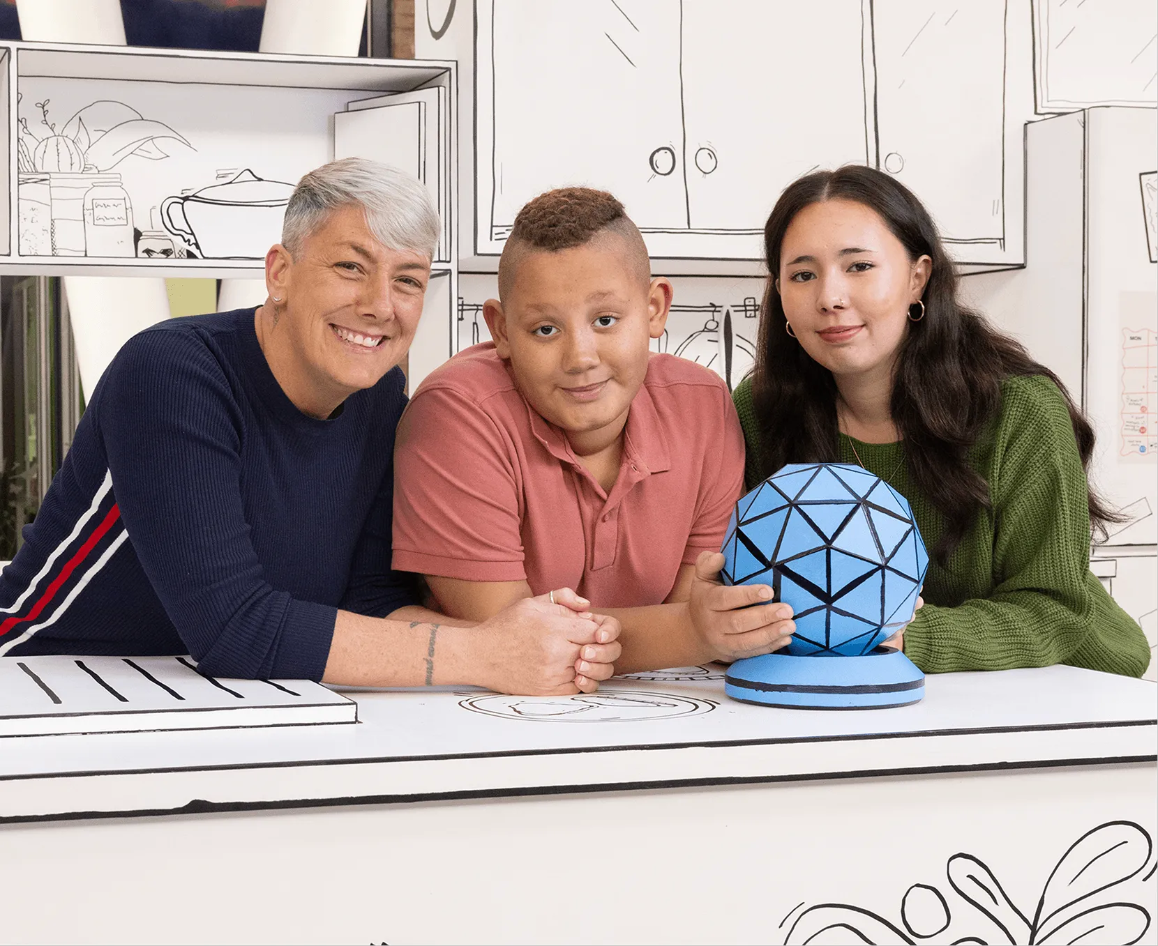 Three people smiling and leaning on a white table with black line drawings, one holding a blue geometric sphere.