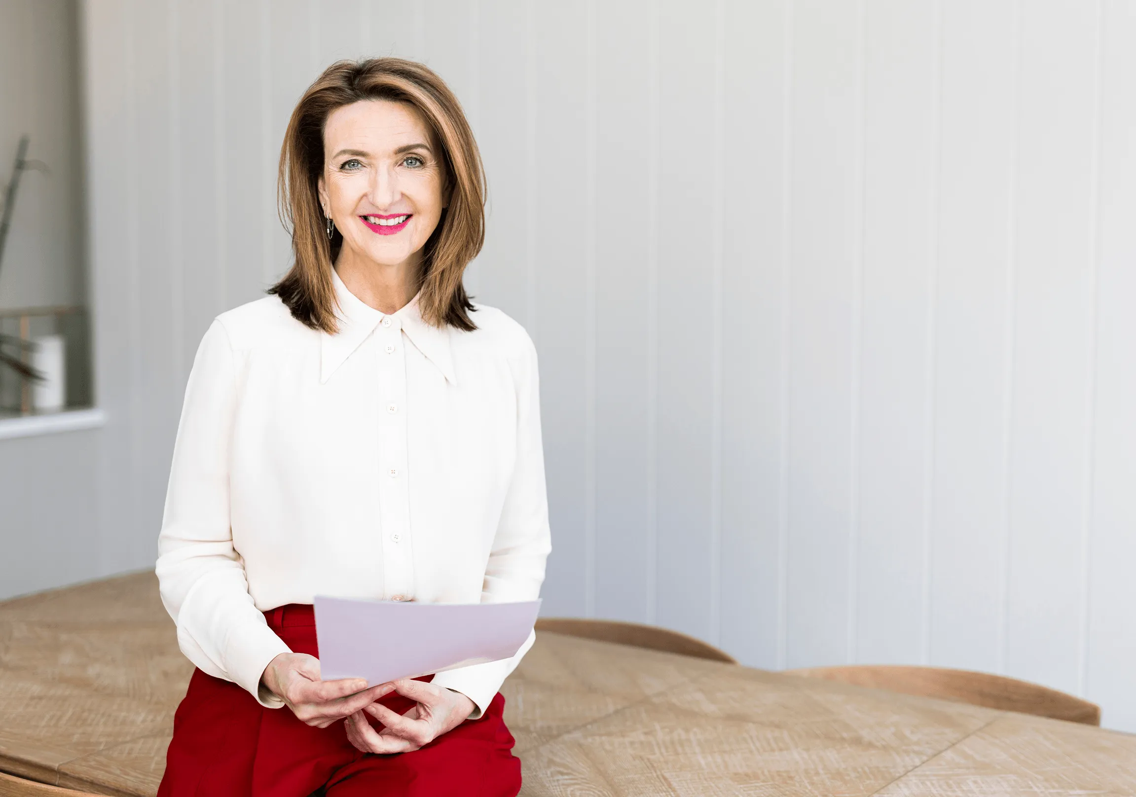 Smiling woman in a white blouse and red pants seated on a wooden table holding a sheet of paper.