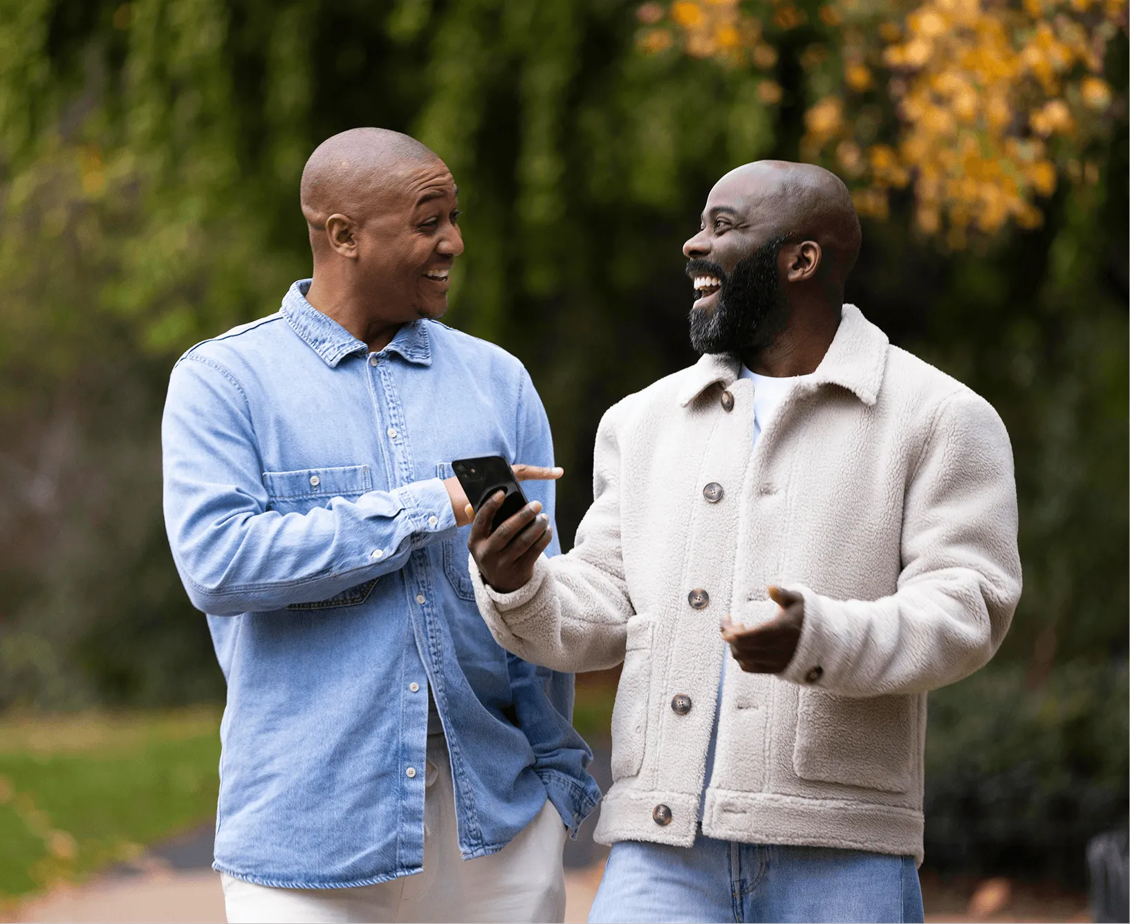 Two men smiling and conversing outdoors, one showing something on his smartphone to the other.