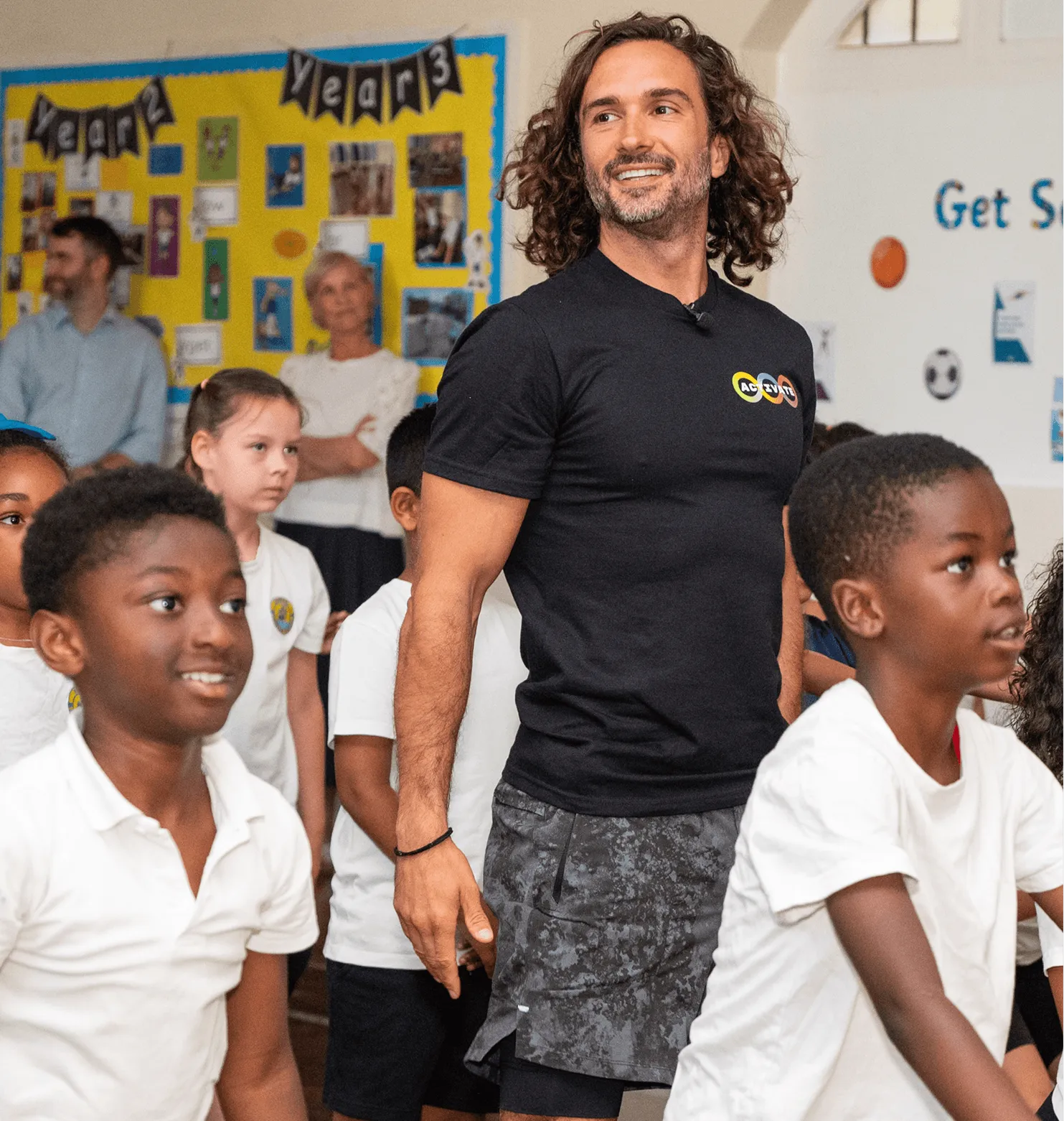 Joe Wicks with curly hair wearing a black t-shirt stands among children in white shirts in a classroom with a Year 2 and Year 3 bulletin board in the background.