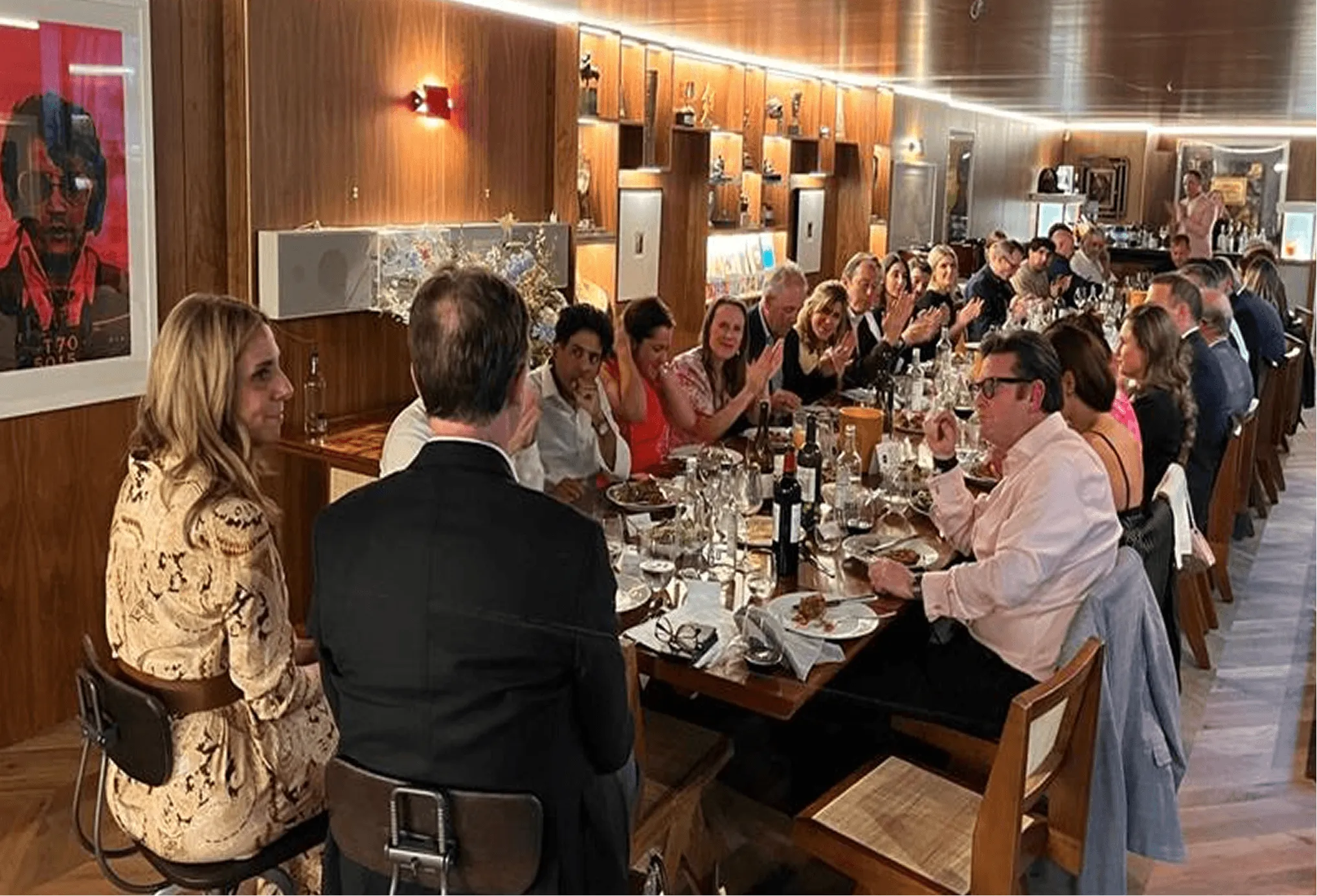 Large group of well-dressed people seated at a long dining table in a warmly lit restaurant, enjoying a meal and conversation.