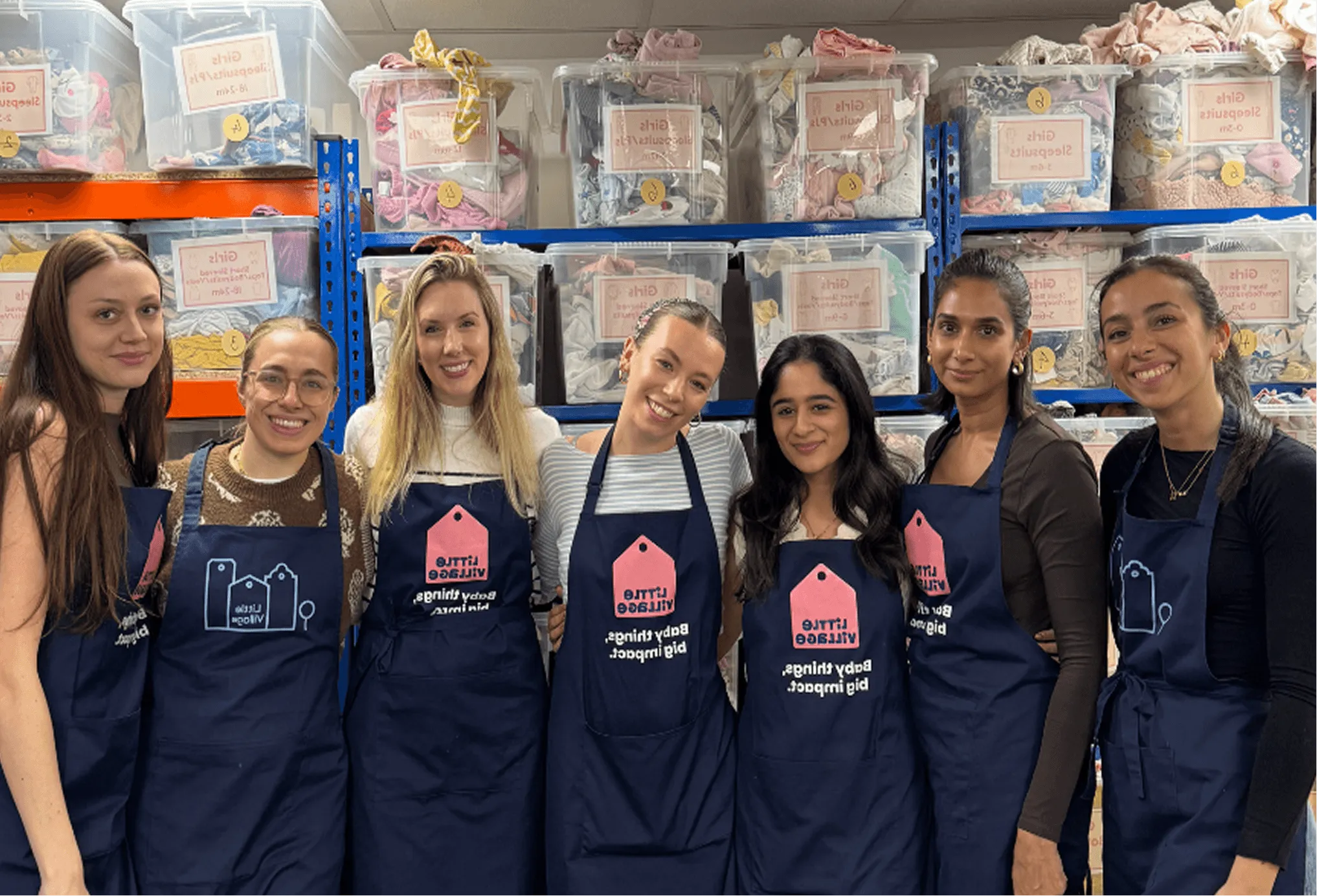 Seven women standing close together wearing navy aprons with ‘Little Village’ logos in front of shelves filled with labeled plastic bins of baby clothes.