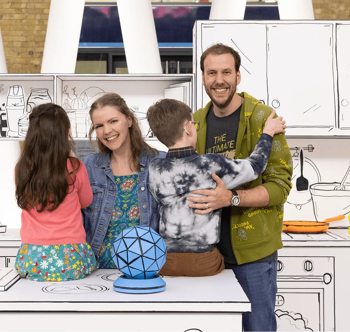 Happy family of four posing in a kitchen with cartoon-style black and white drawn cabinets and appliances in the background.