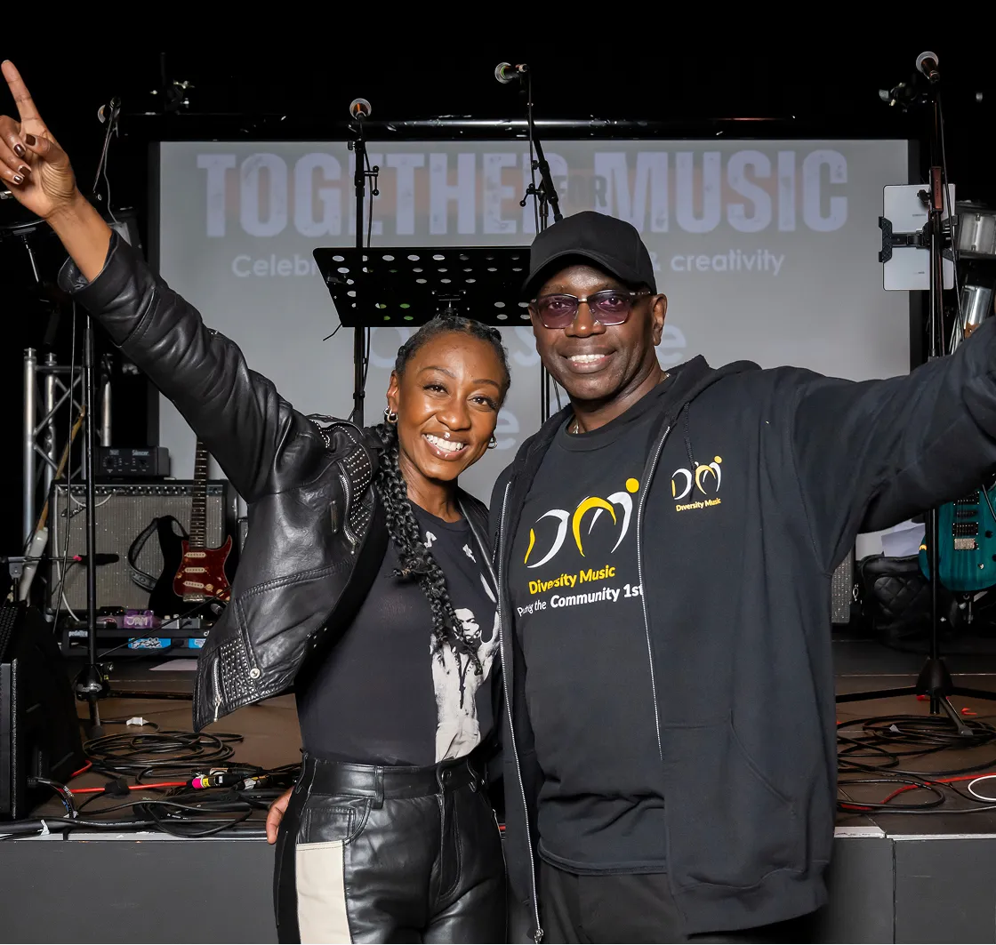 Two smiling people with arms raised posing in front of musical equipment and a stage backdrop reading 'Together Music'.